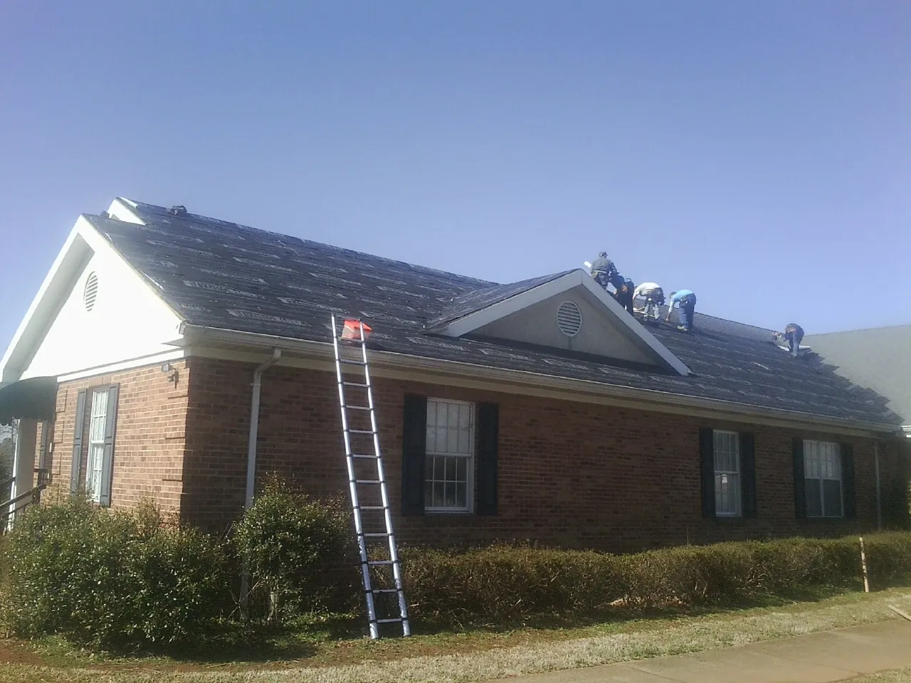 Crew tearing shingles off a brick ranch roof with a ladder set — Emergency Roof Repair work by our crew in Cat Spring